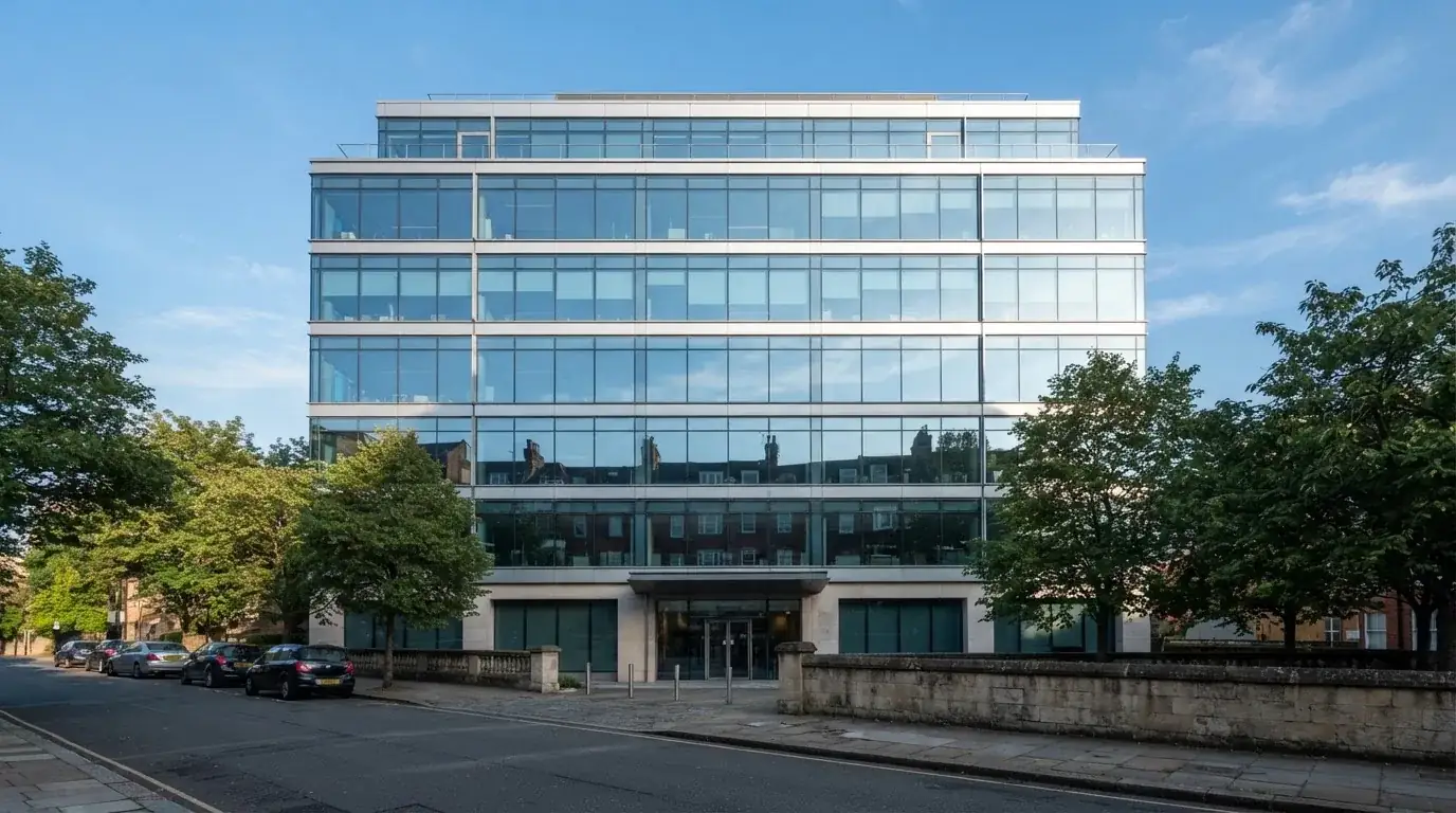 Un edificio de oficinas moderno con cristales reflectantes bajo un cielo despejado, visto desde una calle tranquila.
